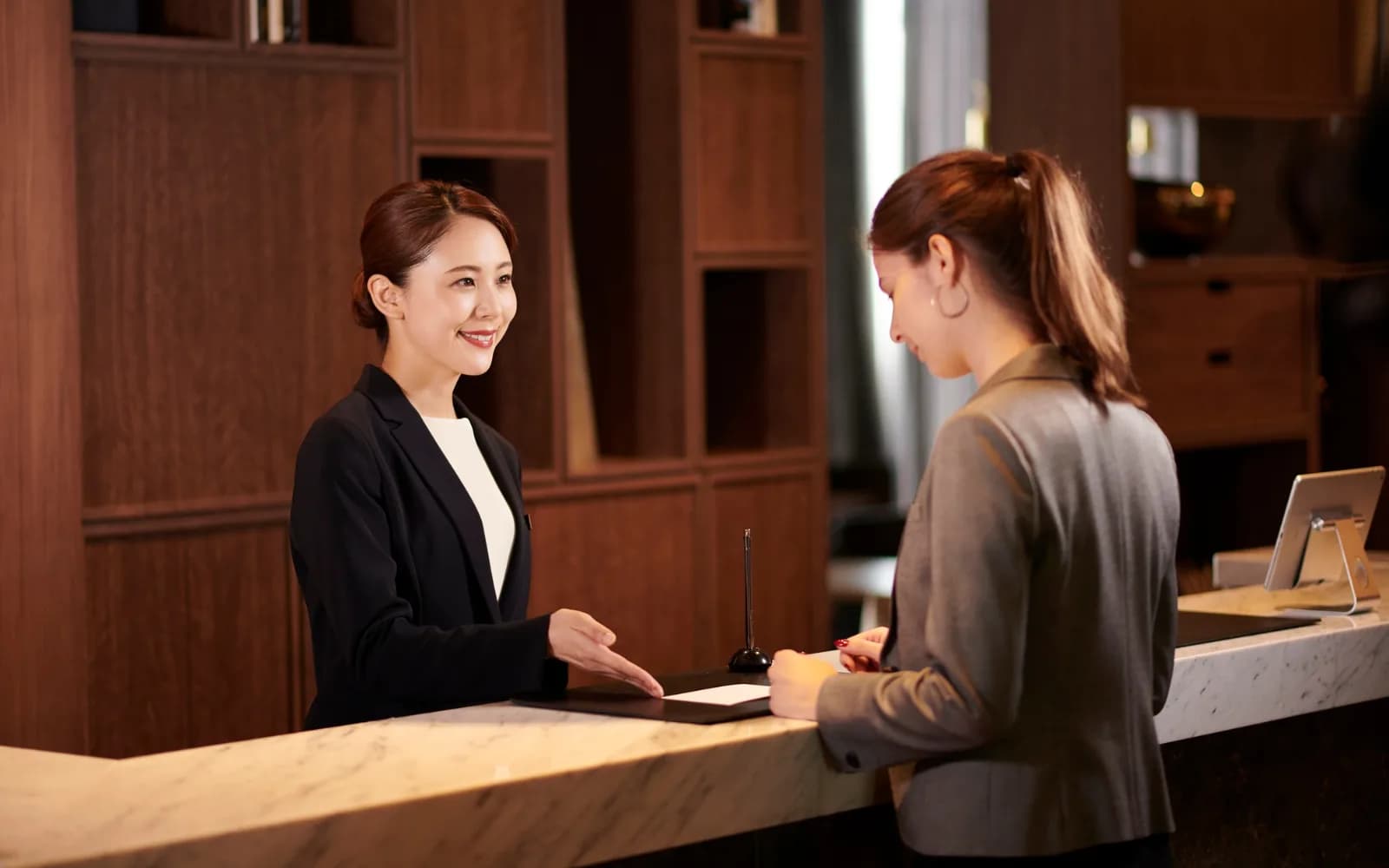 Hotel receptionist welcoming a guest at a marble front desk in a warm wood-panelled lobby