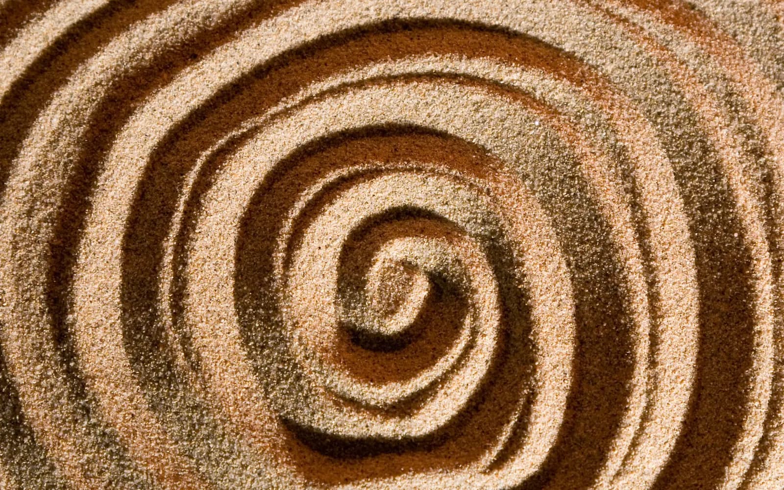 Spiral traced in fine sand, each ring measured and deliberate, circling toward a precise centre